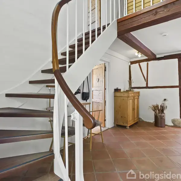 En elegant, buet trappe med trægelænder svæver opad. Den står i en lys gang med terrakottafliser, synlige bjælker og møbler som et skab og et bord med en plante.