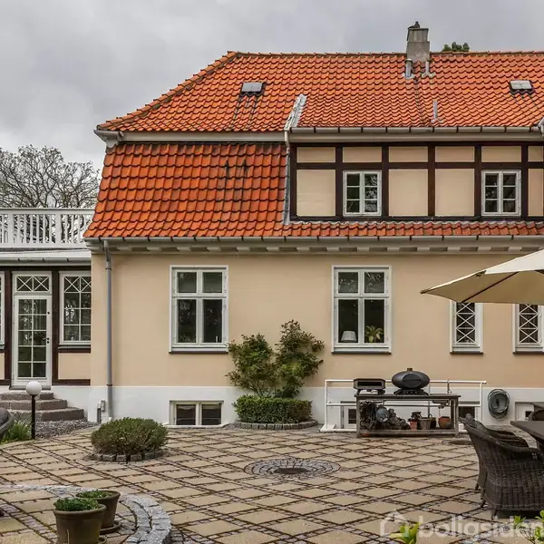 Et hus med rødt tegltag står i en baggård. Der er bord og stole samt en parasol på en flisebelagt terrasse. Skyer dækker himlen.