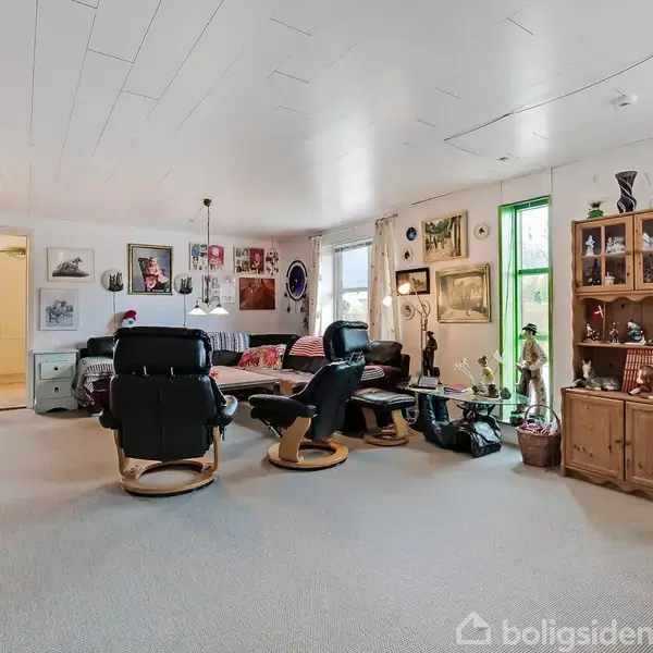 Stue med tre sorte lænestole omkring et rundt sofabord. Vægge dekoreret med billeder. Hylder og skabe med pyntegenstande. Vinduer til højre med lysindfald.