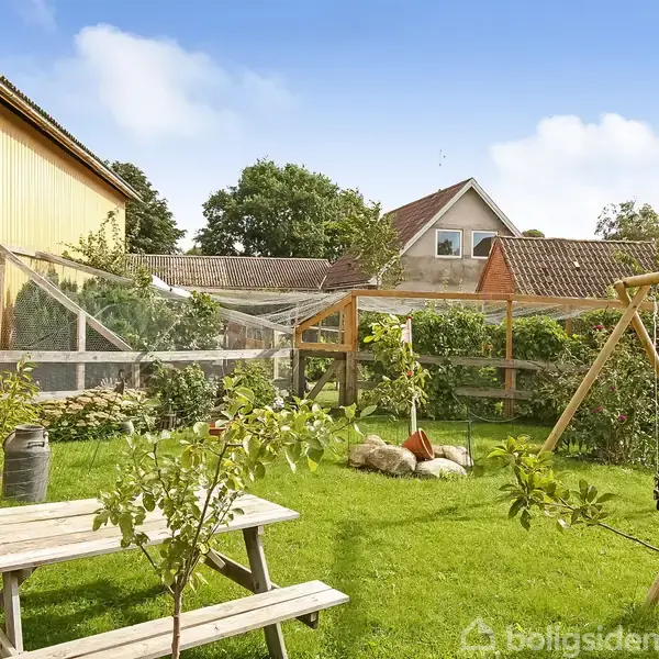 Et bord-bænk-sæt står på en græsplæne ved siden af en gyngestativ. I baggrunden ses et drivhus ved siden af en gul bygning under en klar blå himmel.
