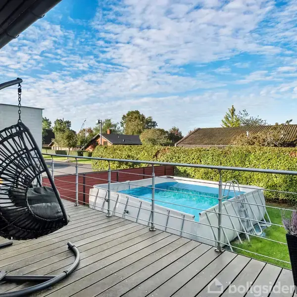 En hængekøjestol hænger stille på en træterrasse. I baggrunden ses en rektangulær pool og en velholdt have, omgivet af hække og forskellige træer under en delvist skyet himmel.