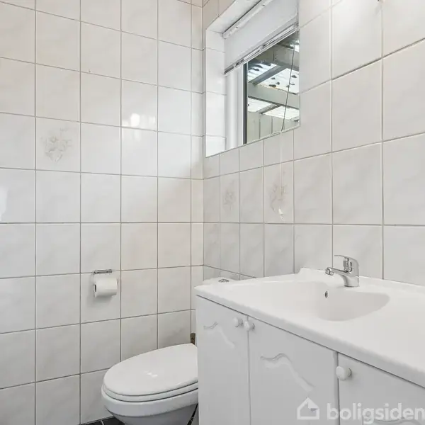 Toilet og håndvask står ved siden af hinanden i et hvidkaklet badeværelse. Rulleholder monteret på væggen. Vindue over spejl giver naturligt lys.