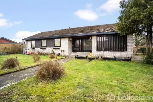 En hvid murstensbungalow med brune vinduer står på en græsplæne, omkranset af en have med buske og en flisebelagt gangsti fører op til hoveddøren under en blå himmel.