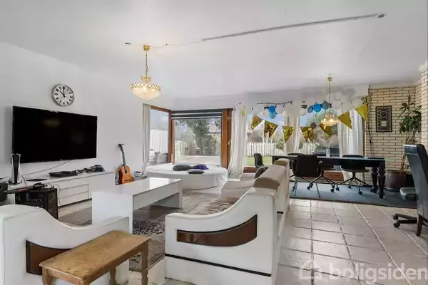 En stue med en hvid sofa står foran et stort fladskærms-tv på væggen. Et sofabord står midt i rummet, dekoreret med en guitarlignende genstand ved siden af. Store vinduer lukker lys ind, og et skrivebord med kontorstole er i baggrunden. Farverige bannere hænger fra loftet.