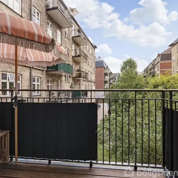 Balkon med sort gelænder, der har en farverig parasol og bænk, vender ud mod grønt område. Omgivet af murstensbygninger med flere altaner i baggrunden.