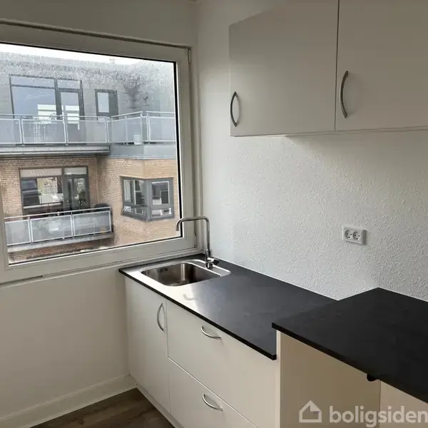 Et lille køkken med hvidt skabsinventar. En vask med en enkelt hane er indbygget i en sort bordplade. Gennem vinduet ses et moderne bygningskompleks med altaner.