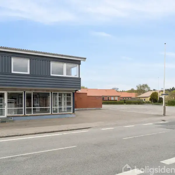 Toetagers bygning med store vinduer i nederste etage, står forladt ved en stille vej. Omkringliggende område inkluderer parkeringsplads og boliger med røde tage under en blå himmel.