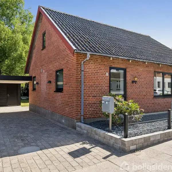 En rød murstenshus med skråt tag står i solskin. Huset har en indkørsel og carport til venstre. Foran huset er der en lille have med buske.