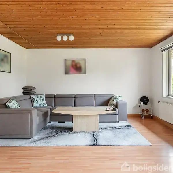 Gråt sofahjørne står i en stue med trægulv og træloft. Et lyst sofabord er placeret foran sofaen. Væggene har tre billeder, og et stort vindue giver lys.