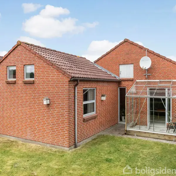 Rødt murstenshus med et glastilbygget udestueområde, omgivet af en velholdt græsplæne og grøn hæk. To paraboler er monteret på taget. Himlen er let skyet.