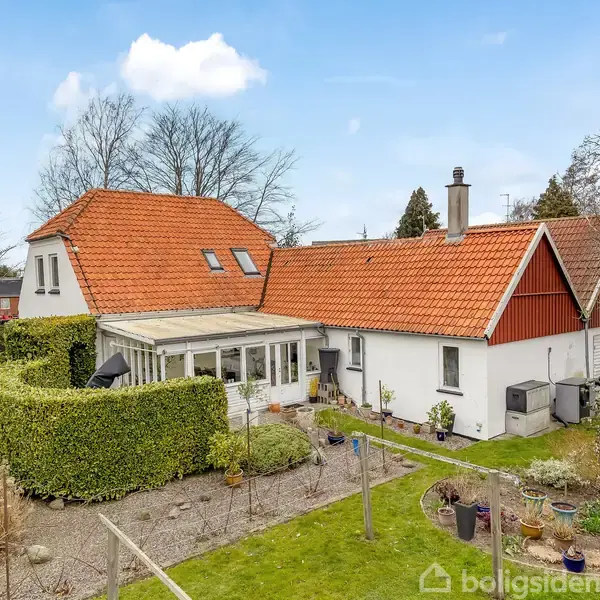 Hus med rødt tegltag og hvid facade, omgivet af have med buske og træer, under blå himmel. Terrasse med glasoverdækning langs siden. Græsplæne med krukker og planter omkring.