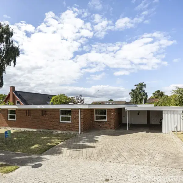 En rød murstensvilla står stille i et boligområde, omgivet af grønne træer og en brolagt indkørsel. Huset har en carport på siden og en blå himmel ovenover.