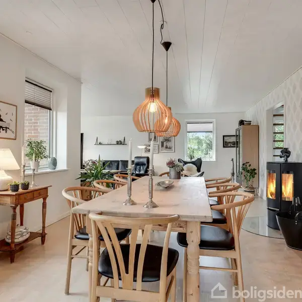 Et spisebord med træstole står i en lyse, indbydende spisestue. Over bordet hænger lamper, og en brændeovn er tændt ved siden af planter og hylder.