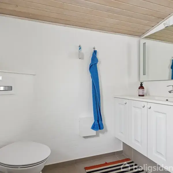 Et moderne badeværelse indeholder et væghængt toilet, en blå håndklæde på væggen og et skab med spejl, der reflekterer lyset. Rummet er lyst med hvidmalede vægge.