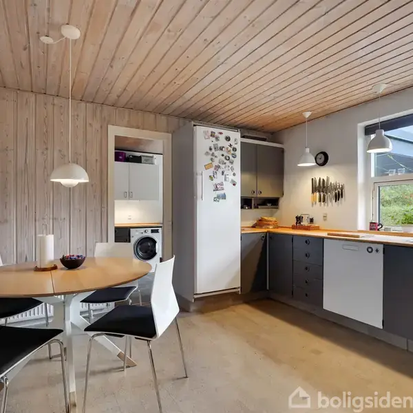 Køkken med spisebord og fire stole. Bordet har en skål og en lysestage. Vægge og loft i træ. Køleskab med magneter. Køkkenvask og vindue med udsigt til haven.