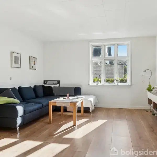 Hus med sort tag og røde mursten står foran en stor græsplæne. Til venstre er et drivhus. En træterrasse ses foran huset.