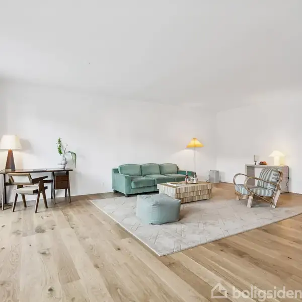 En stue med træbord indeholder en grøn sofa og to stole arrangeret på et tæppe. To lamper lyser op i rummet, der har hvide vægge og trægulve. En vase står i forgrunden.