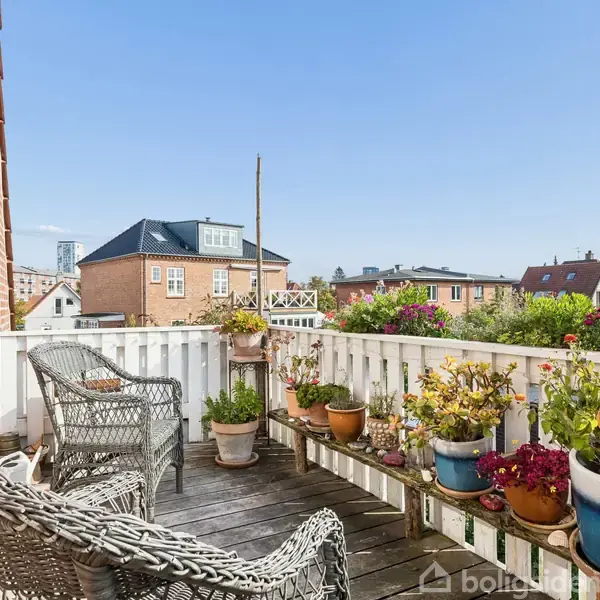 To fletstole står på en altan omringet af blomsterkrukker. Altanen har udsigt over en række hustage under en klar blå himmel.