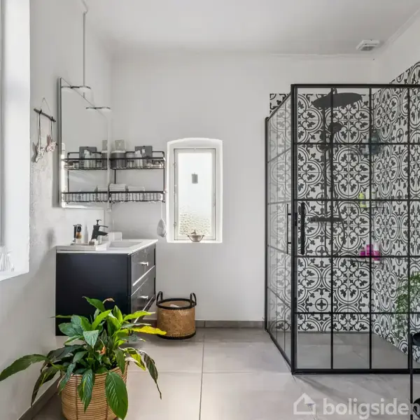 En moderne badeværelseindretning med en sort brusekabine med dekorativt mønster. En sort vaskeskab står ved siden af, omgivet af planter og en stor rude med naturligt lys.