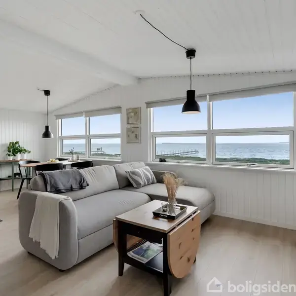 Grå sofa står i lyse stue med stort vindue, der viser udsigt over havet. Rundt om er spisebord og indretning med sort-hvide lamper og lyse træmøbler.