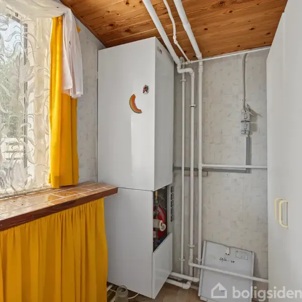 En hvid vaskemaskine står i et lille rum med gule gardiner og et træloft. Maskinen er omgivet af rør, og der er et vindue med blondegardiner til venstre.