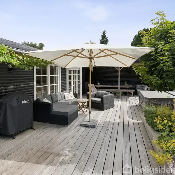 En terrasse med træplanker har en parasol, lounge-sæt og gasgrill tæt på et sortmalet hus. Havemøblerne er omgivet af grønne planter under en klar himmel.