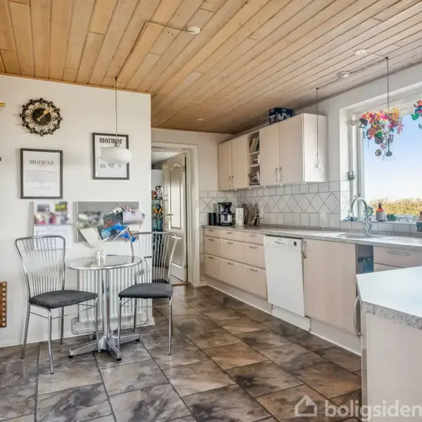 Køkkenet har beige skabe og en marmoreret gulvflise, med et bord og stole placeret midt i rummet. Store vinduer med farverige dekorationer tillader udsigt til det fri.