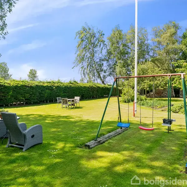 Gyngestativ med to gynger står på en velplejet græsplæne. Liggestole og bord med stole ses i baggrunden, omgivet af grønne træer og buske på en solrig dag.