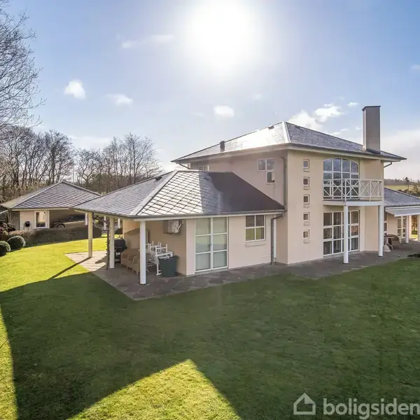 En stor, moderne villa står på en velplejet græsplæne omgivet af træer og åbne marker. Solen skinner ovenfra, skaber skygger og giver en lys atmosfære.