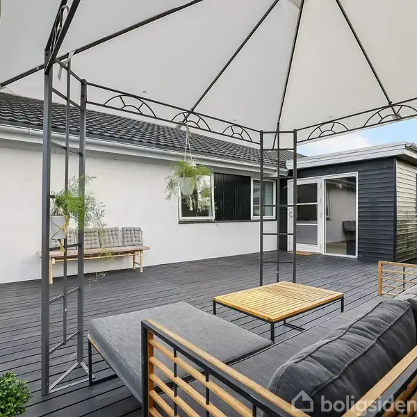 En terrasse med en pavillon, hvor der står et sofaarrangement med bord. I baggrunden ses en hvid murstensbygning og en sort skur, omgivet af grønne planter.