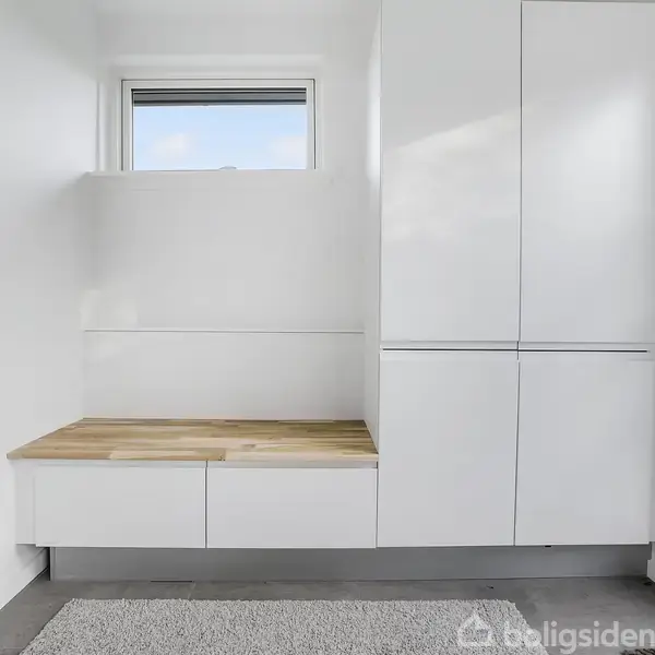 Hvidt skab monteret på væggen står ved siden af en indbygget bænk med trægulv. Bænken er under et vindue og på en lys tæppebelagt gulv.
