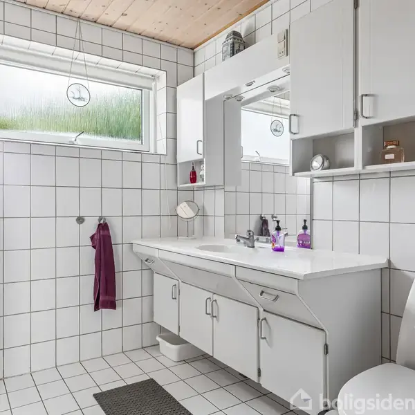 Badeværelse med hvidt flisegulv og -vægge. Toilettet er til højre, og der er et dobbelt vaskeskab med spejl og en hylde over. Et vindue giver naturligt lys.