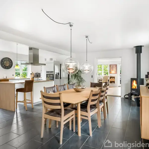 Spisebord med otte stole står i et lyst, åbent køkkenalrum, hvor en brændeovn i hjørnet afgiver varme. Store vinduer og moderne lamper skaber en indbydende atmosfære.