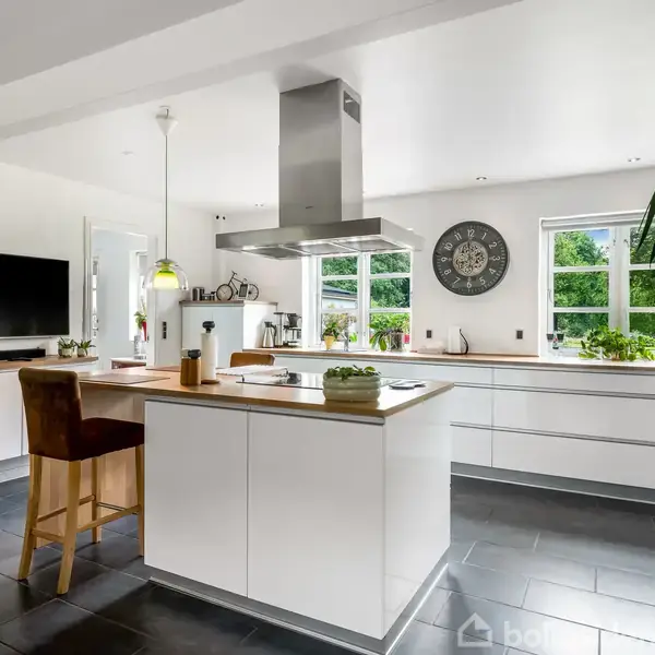 Et moderne køkken med hvide skabe og en central ø med barstol. Rummet er lyst med store vinduer, grønne planter og et vægur på væggen.