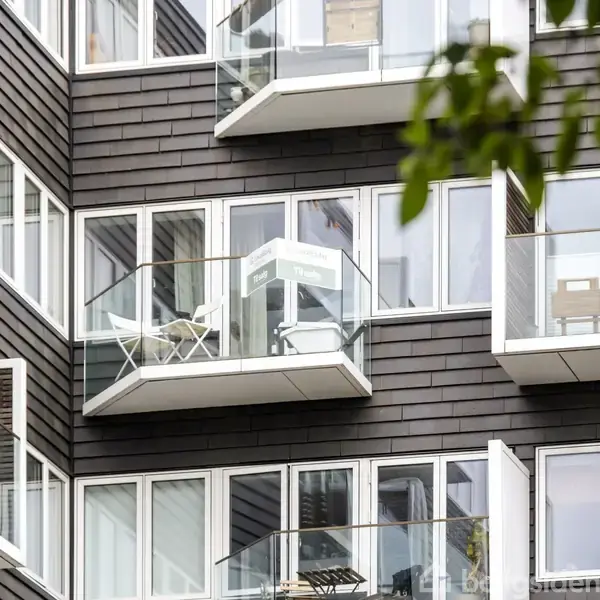 Et lejlighedskompleks med mange altaner er hovedfokus. Bygningen er moderne med glas- og træfacader. Det er dagtid, og himlen er klar.