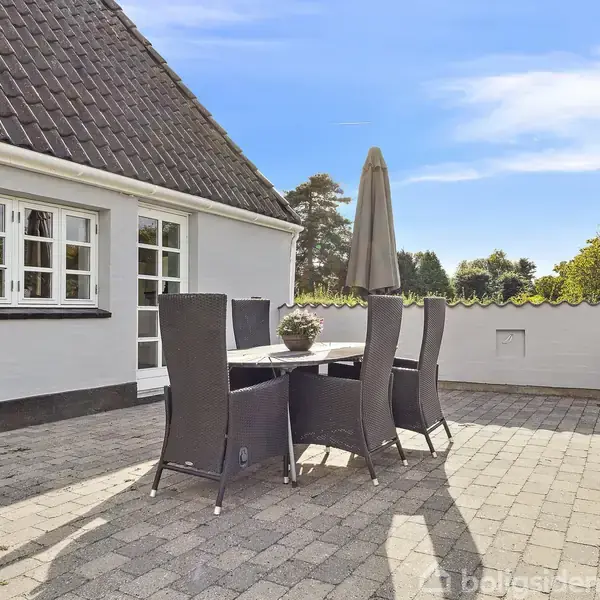 En udendørs terrasse med et havebord og seks stabelbare stole. Bordet har en dekorativ krukke. Solrig dag med blå himmel og bygning i baggrunden.