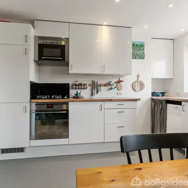 Et moderne køkken med hvide skabe og træbordplader. En ovn og mikrobølgeovn er indbygget i skabene. Et lille spisebord og stol står i forgrunden nær en blomsterskål.