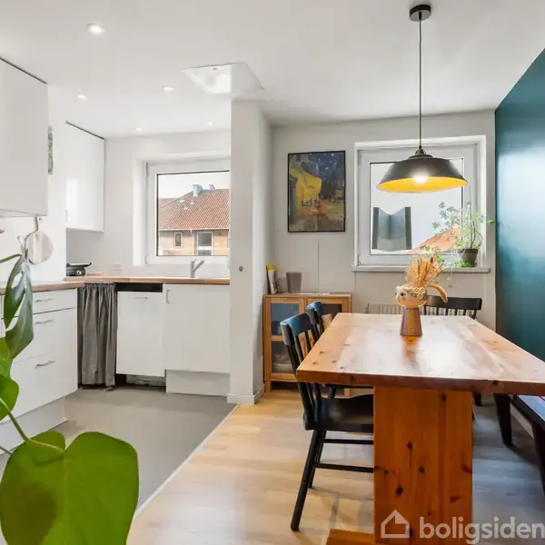 Et lyst køkken-alrum med spisebord af træ flankeret af stole og bænk, placeret nær en stor, grøn plante. Køkkenet har hvide skabe og gråt gulv med vindue i baggrunden.