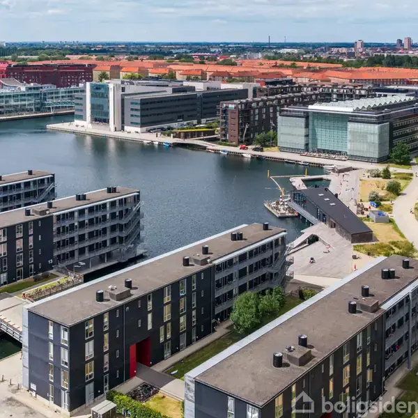 Stue med trægulv i en moderne lejlighed, store vinduer åbner mod en balkon med udsigt over andre bygninger. Rummet er lyst med hvide vægge. Ingen tekst bortset fra "Boligsiden".