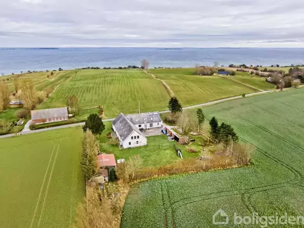 Et hvidt hus med mørkt tag står stille i et åbent landligt område omgivet af grønne marker og træer. En indkørsel fører til huset.