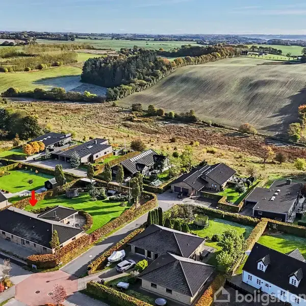 Boligkvarter med huse omgivet af grønne haver. Husene ligger op ad åbne marker og skove i bakkede landskaber. Det er en solrig dag med klar udsigt over området.