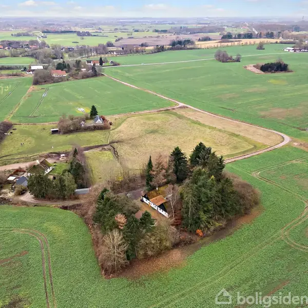 Et hus omgivet af tæt beplantning ligger på en grusvej i et vidtstrakt landskab med marker og spredte træer. Veje og bygninger ses i det fjerne.