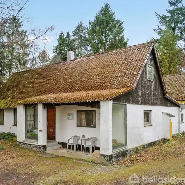 Et hvidt landhus med brunligt tag står i en skovklaring. Foran huset er der et lille udendørs område med et bord og stole.
