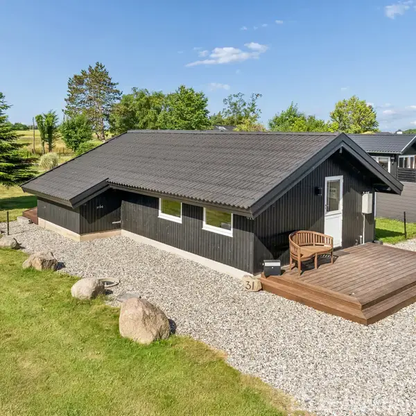 Et sort træt hus med grått tag står på en grusbelagt grund, omgivet af græs og nogle træer. En trækæde og stentryk dekorerer området.