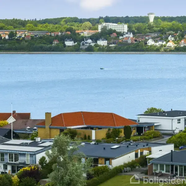 Boliger med flade tage og røde tage ligger stille ved en søbred. På den anden side af søen ligger flere huse og en by omgivet af grønt landskab og skov.