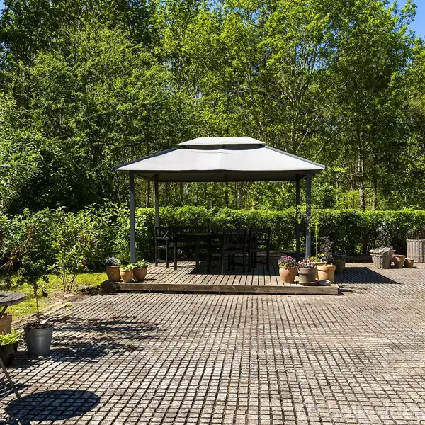 Pavillon står på en flisebelagt terrasse med flere potteplanter omkring. Omgivet af frodige grønne træer og buske, skaber det en afslappet udendørs atmosfære. Ingen tekst er til stede.