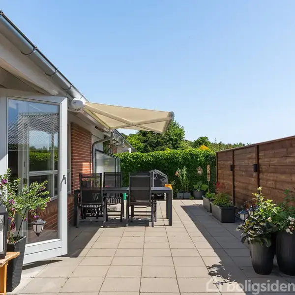 Terrasse med flisebelægning, hvor et bord med stole står under en åben markise. Til højre står plantekrukker langs et hegn, og til venstre er der murstensvæg med en dør.