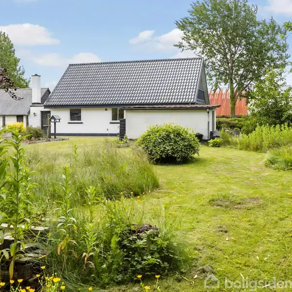 Et hvidt hus med sort tag står i en frodig have med højt græs og blomster. Der er træer i baggrunden og en klar blå himmel.