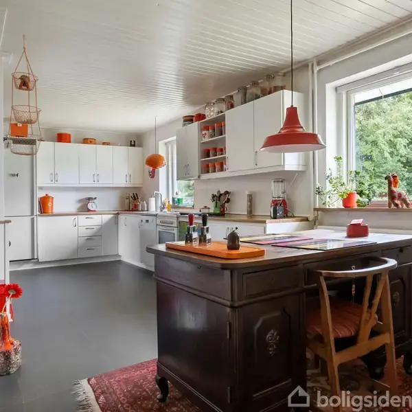 Et træbord med stol står i lys køkkenindretning. Der er planter og røde lamper ved et stort vindue. På hylder står køkkenredskaber og orange dekorationer skaber livlig atmosfære.