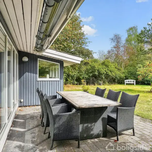 En udendørs spisebordssæt med otte sorte kurvestole står på en terrasse ved siden af et blågråt hus. Omkring terrassen er der en grøn have med træer og buske.